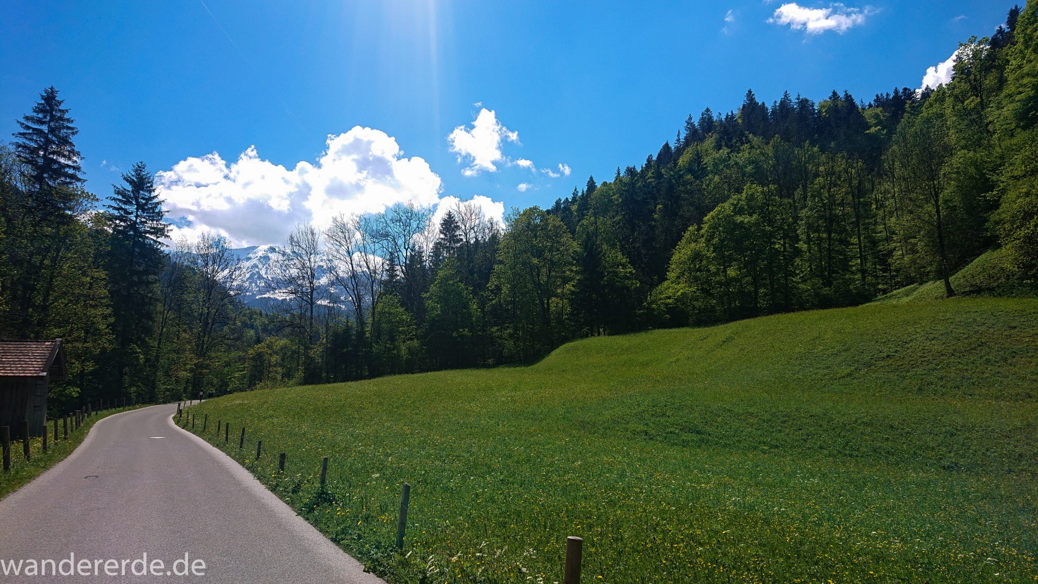 Rundwanderung Partnachklamm im Wettersteingebirge bei Garmisch-Partenkirchen, Bayern, Wanderweg zunächst über asphaltierte Straße, aber ohne Autos, saftig grüne Wiesen und schöner Wald ringsum, in der Ferne Blick auf schneebedeckte Berge