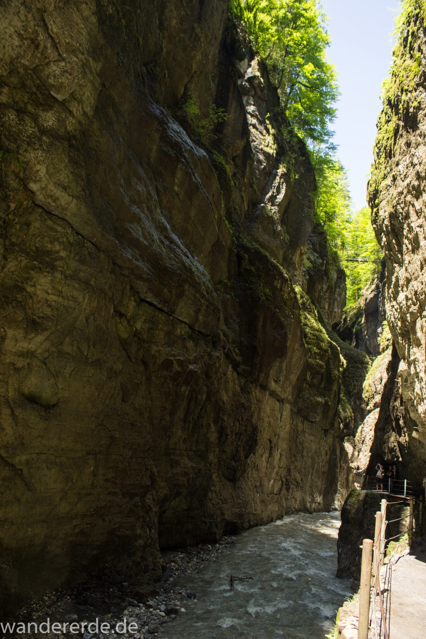 Rundwanderung Partnachklamm im Wettersteingebirge bei Garmisch-Partenkirchen, Bayern, steil aufragende Felswände, Licht strahlt von oben in die Klamm, oben wachsen Bäume, kühl und nass, Sicht auf eiserne Brücke von der man Aussicht in die Klamm hat, gesicherter Wanderweg, Fluss Partnach bahnt sich Weg durch die Klamm