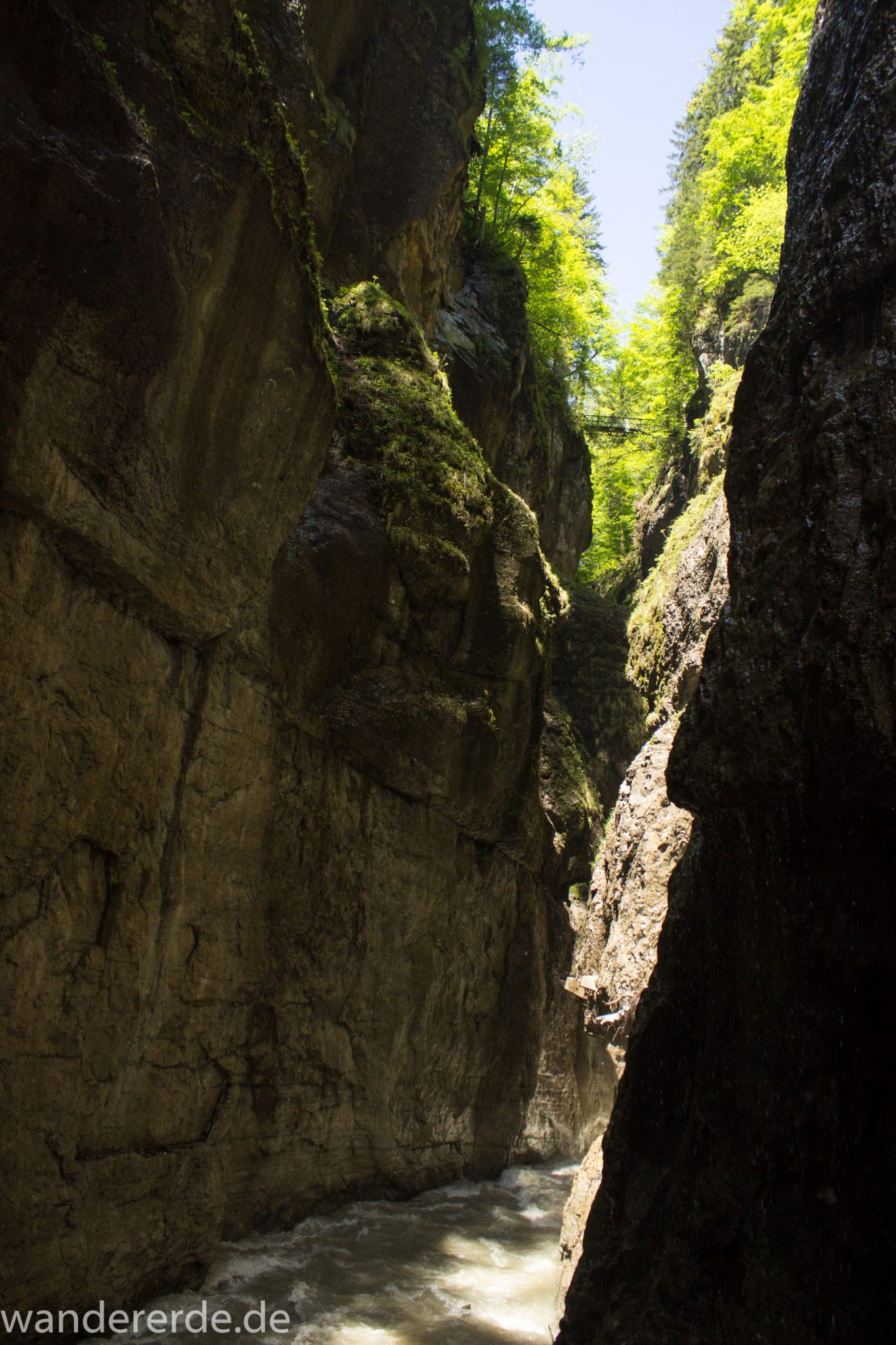 Rundwanderung Partnachklamm im Wettersteingebirge bei Garmisch-Partenkirchen, Bayern, steil aufragende Felswände, Licht strahlt von oben in die Klamm, oben wachsen Bäume, kühl und nass, Sicht auf eiserne Brücke von der man Aussicht in die Klamm hat