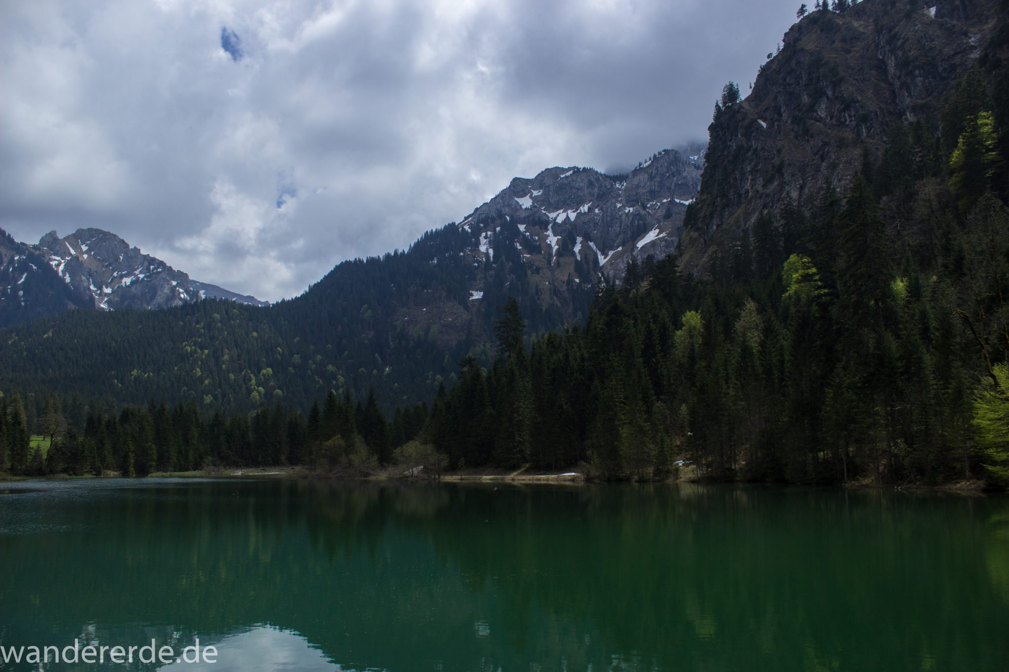 Wanderung zur Kenzenhütte in den Ammergauer Alpen, Aussicht beim Bockstallsee auf die Berge, teils schneebedeckte Gipfel in Wolken gehüllt, Frühjahr in den bayerischen Alpen, dichter grüner Wald umgibt den Bockstallsee