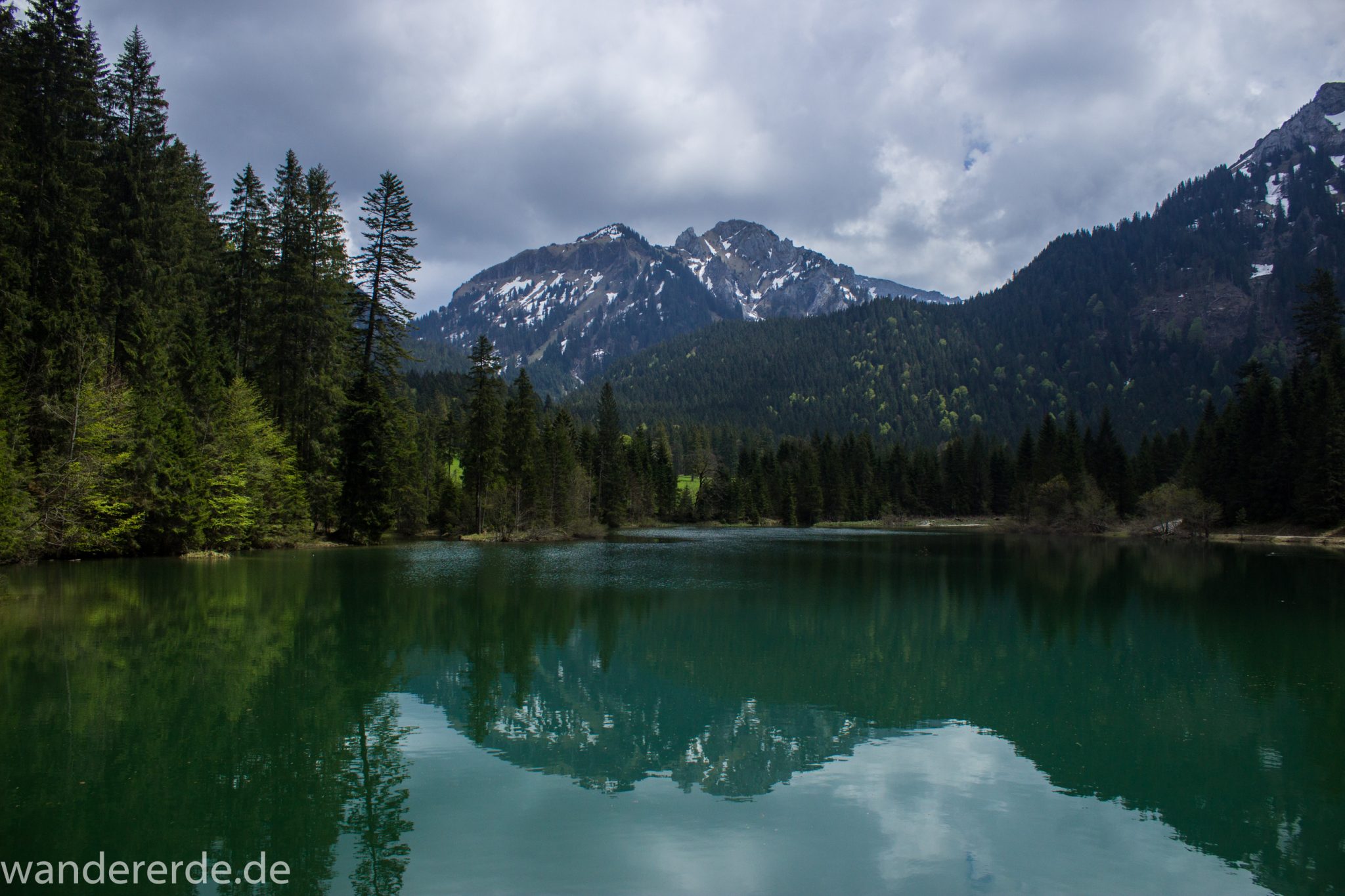 Wanderung zur Kenzenhütte in den Ammergauer Alpen, Aussicht beim Bockstallsee auf die Berge, teils schneebedeckte Gipfel in Wolken gehüllt, Frühjahr in den bayerischen Alpen, dichter grüner Wald umgibt den Bockstallsee