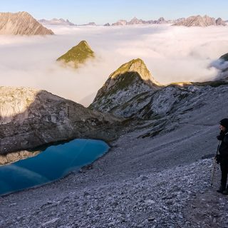 Alpenüberquerung Fernwanderweg E5 Oberstdorf Meran, 3. Etappe von Memminger Hütte zur Skihütte Zams, E5 Wanderweg führt zu Beginn der dritten Etappe ab der Memminger Hütte über steilen und ausgesetzten Wanderweg über die Seescharte, Wanderer blickt auf kristallklaren Bergsee während Aufstieg zur Seescharte auf sehr schmalen Wanderweg über viel Geröll, Aussicht auf Berge der Alpen die in Nebel gehüllt sind