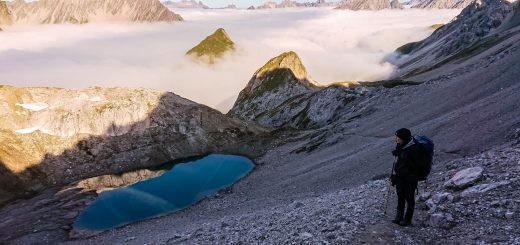 Alpenüberquerung Fernwanderweg E5 Oberstdorf Meran, 3. Etappe von Memminger Hütte zur Skihütte Zams, E5 Wanderweg führt zu Beginn der dritten Etappe ab der Memminger Hütte über steilen und ausgesetzten Wanderweg über die Seescharte, Wanderer blickt auf kristallklaren Bergsee während Aufstieg zur Seescharte auf sehr schmalen Wanderweg über viel Geröll, Aussicht auf Berge der Alpen die in Nebel gehüllt sind