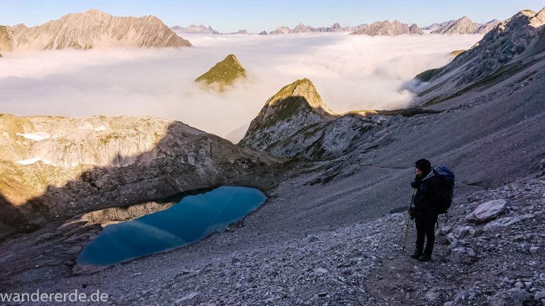 Alpenüberquerung Fernwanderweg E5 Oberstdorf Meran, 3. Etappe von Memminger Hütte zur Skihütte Zams, E5 Wanderweg führt zu Beginn der dritten Etappe ab der Memminger Hütte über steilen und ausgesetzten Wanderweg über die Seescharte, Wanderer blickt auf kristallklaren Bergsee während Aufstieg zur Seescharte auf sehr schmalen Wanderweg über viel Geröll, Aussicht auf Berge der Alpen die in Nebel gehüllt sind