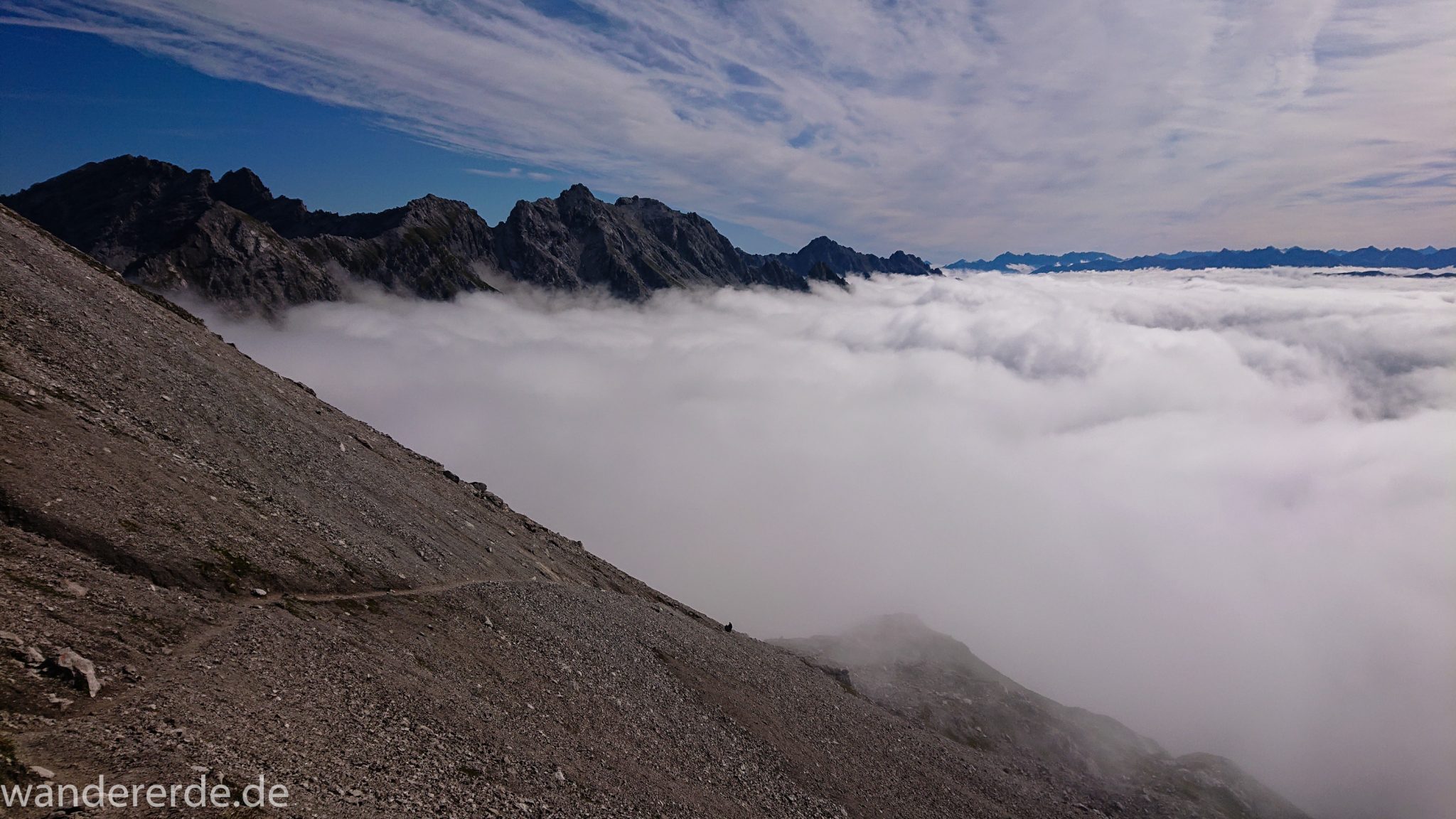 Alpenüberquerung Fernwanderweg E5 Oberstdorf Meran, 3. Etappe von Memminger Hütte zur Skihütte Zams, Blick auf die vor einem liegenden Berge der Alpen bei Erreichen der Seescharte, nach langem, steilen Aufstieg auf der schattigen Seite erreicht man damit die sonnige Seite und somit den Beginn eines lange währenden Abstiegs, Blick auf schmalen Wanderpfad mit viel Geröll während Abstieg ins Lochbachtal Richtung Zams, Berge liegen im Nebel verborgen
