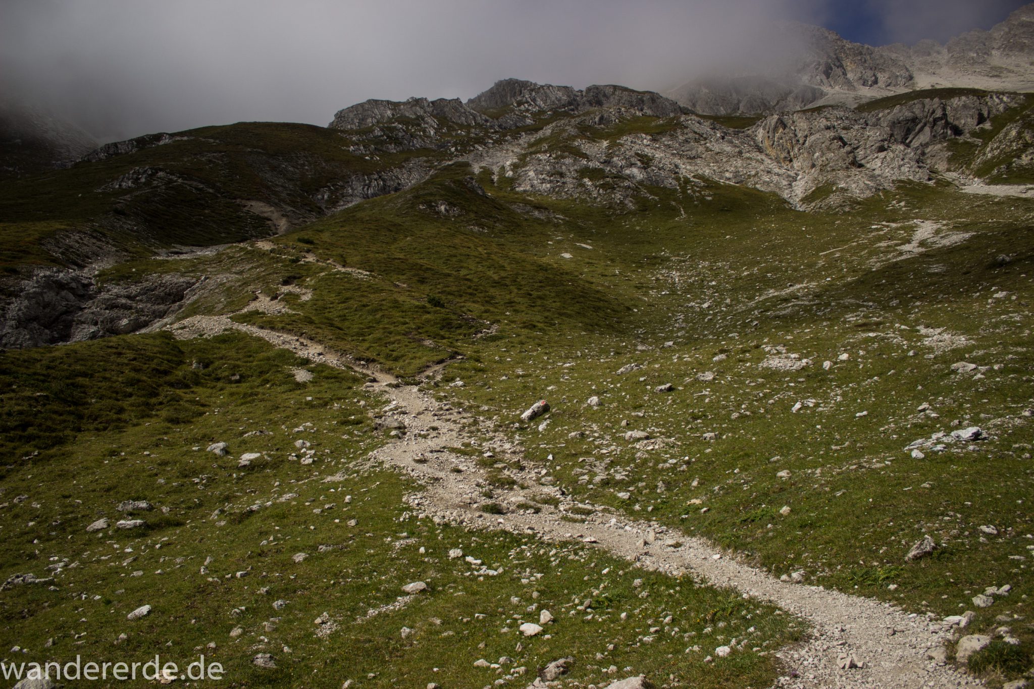 Alpenüberquerung Fernwanderweg E5 Oberstdorf Meran, 3. Etappe von Memminger Hütte zur Skihütte Zams, nach Erreichen der Seescharte folgt ein sehr langer Abstieg ins Lochbachtal Richtung Zams, Aussicht auf Berge und Bergwiesen, Berge und Umgebung liegen teils im Nebel verborgen, Blick auf schmalen Wanderpfad mit viel Geröll