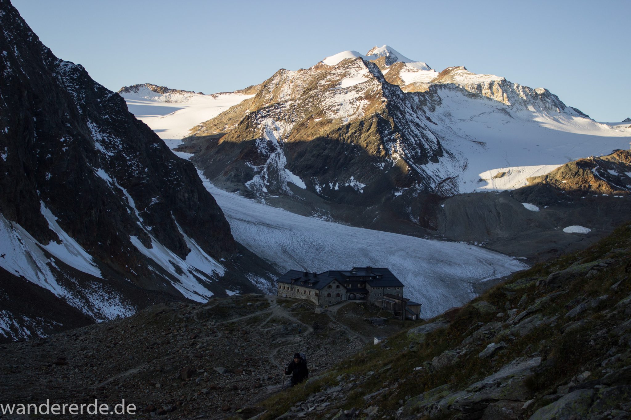 Alpenüberquerung Fernwanderweg E5 Oberstdorf Meran, 5. Etappe von Braunschweiger Hütte zur Martin-Busch-Hütte, zu Beginn der 5. Etappe Blick zurück auf die Braunschweiger Hütte und die umliegenden Berge der Alpen, Wanderer auf steilem und schmalem Wanderweg mit viel Geröll hinauf zum Pitztaler Jöchl, beeindruckende Berge türmen sich vor einem auf, Reste von Schneefeldern