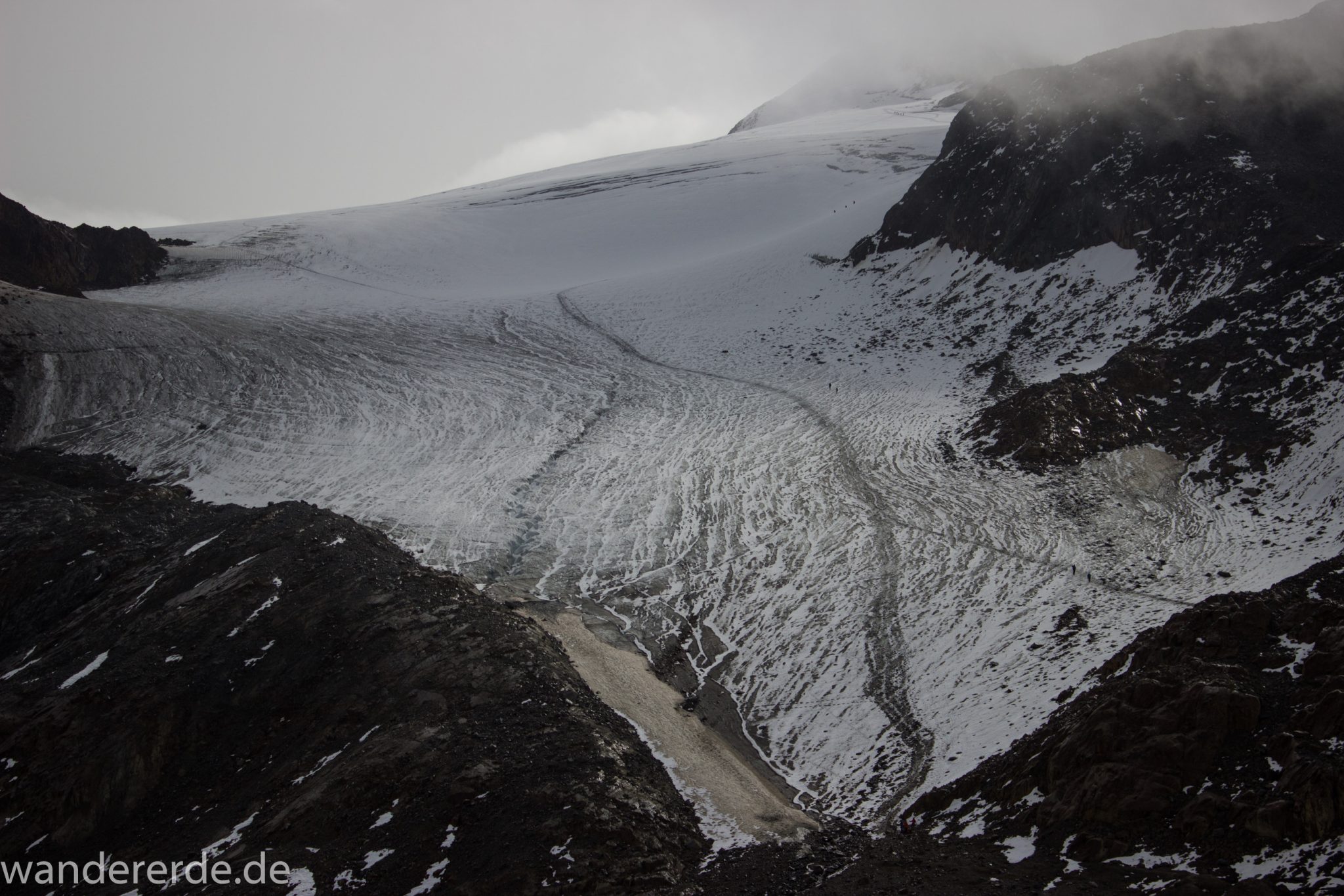 Alpenüberquerung Fernwanderweg E5 Oberstdorf Meran, 6. Etappe von Martin-Busch-Hütte nach Vernagt / Meran, die letzte Etappe der Alpenüberquerung auf dem E5 startet mit dem Aufstieg zur Similaun-Hütte, dem höchsten Punkt der gesamten Alpenüberquerung, umliegende sehr beeindruckende Bergwelt erscheint unwirklich und mystisch, Berghänge mit viel Geröll, Wanderweg verläuft steil bergauf, Blick auf Wanderer die zum Gipfel des Similaun Berges wandern über Schneefelder, nicht Teil des E5