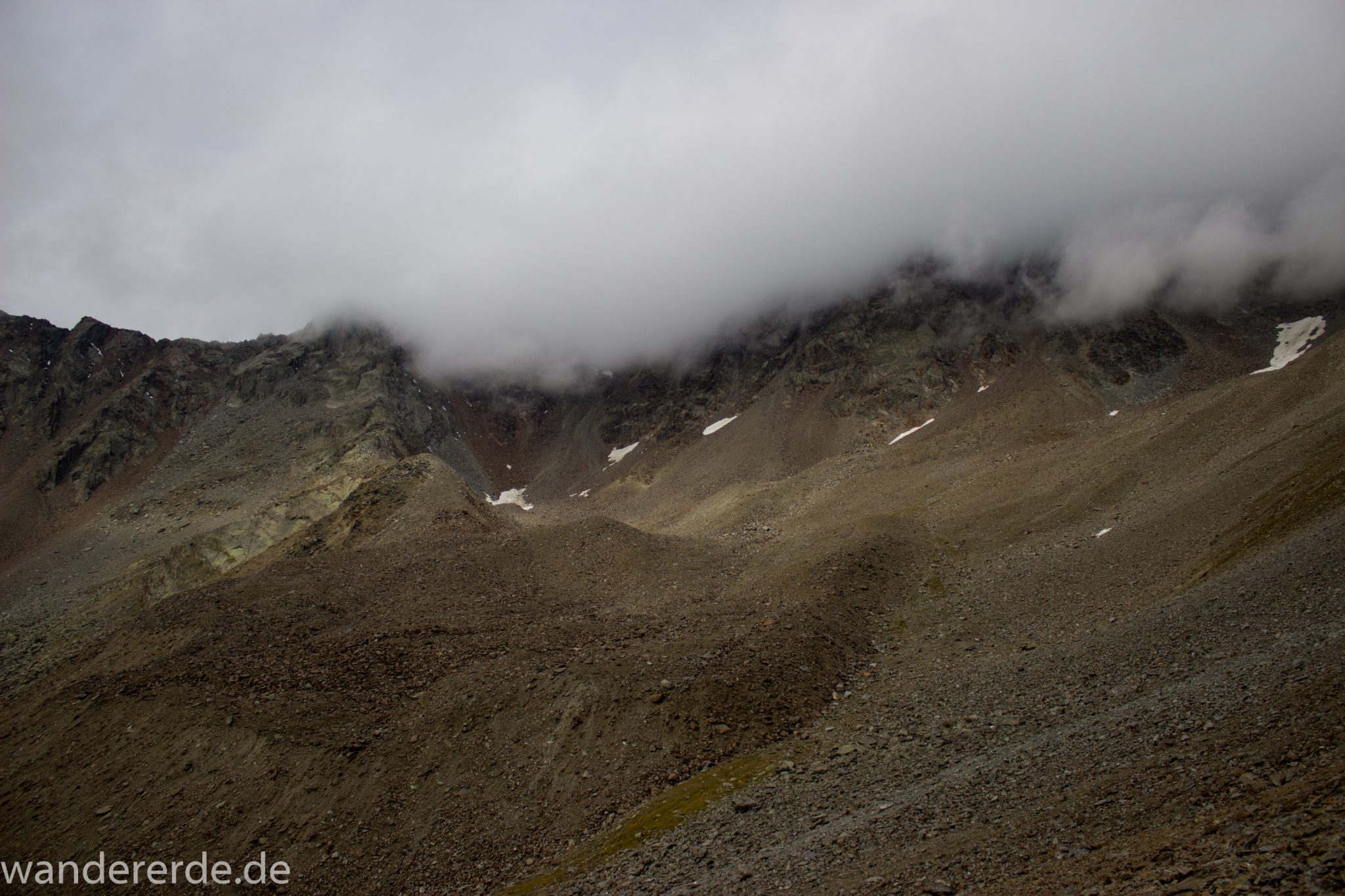 Alpenüberquerung Fernwanderweg E5 Oberstdorf Meran, 6. Etappe von Martin-Busch-Hütte nach Vernagt / Meran, nach dem Aufstieg zur Similaun-Hütte auf der letzten Etappe der Alpenüberquerung auf dem E5 verläuft der Wanderweg steil abwärts Richtung Vernagt im Schnalstal,  Blick auf Berge während Abstieg, erscheint unwirklich und mystisch, Berghänge mit viel Geröll, Wanderung mit steilem Abstieg auf ausgesetztem Pfad, Berggipfel sind in Wolken gehüllt