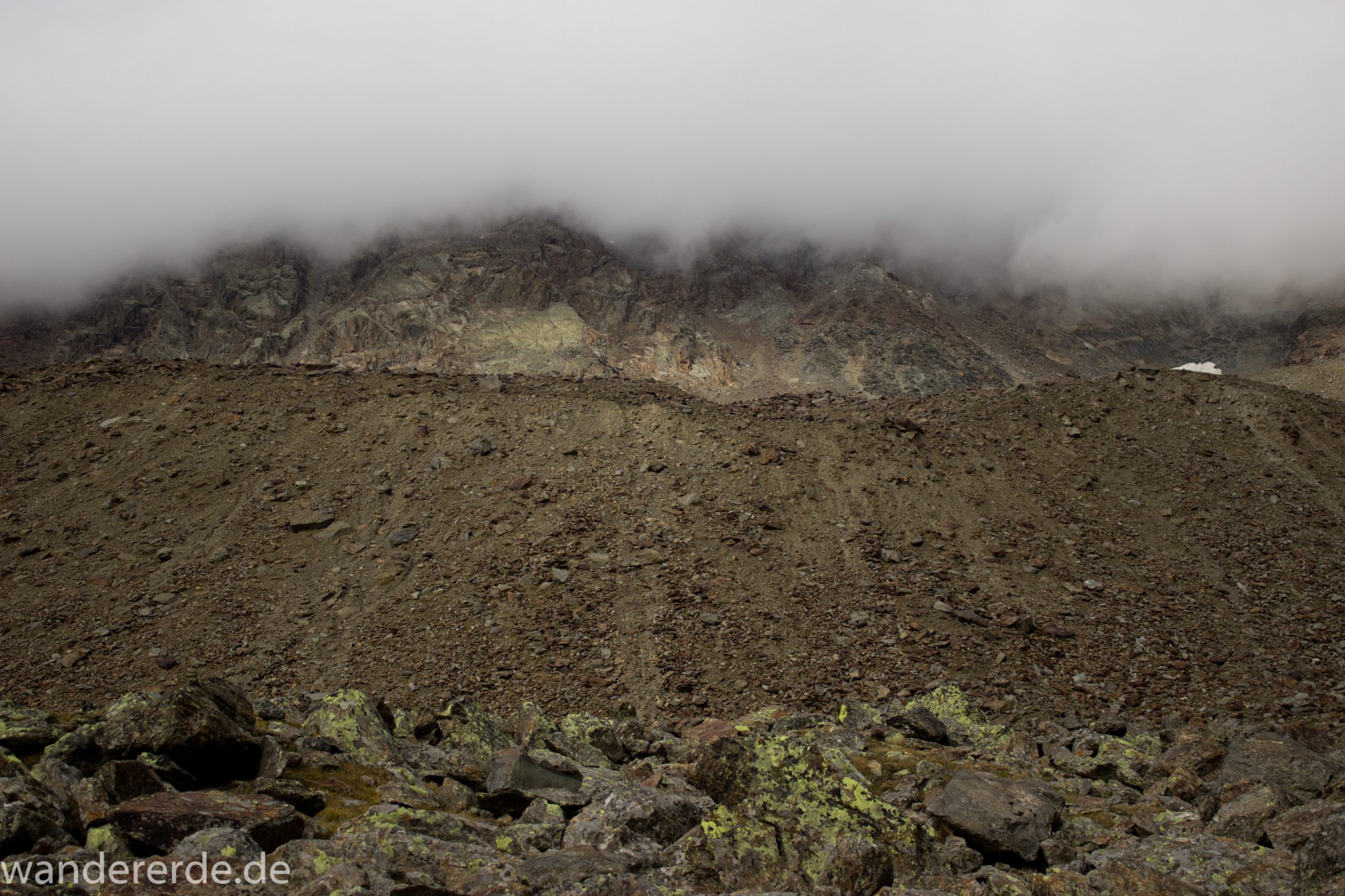 Alpenüberquerung Fernwanderweg E5 Oberstdorf Meran, 6. Etappe von Martin-Busch-Hütte nach Vernagt / Meran, nach dem Aufstieg zur Similaun-Hütte auf der letzten Etappe der Alpenüberquerung auf dem E5 verläuft der Wanderweg steil abwärts Richtung Vernagt im Schnalstal,  Blick auf Berge während Abstieg, erscheint unwirklich und mystisch, Berghänge mit viel Geröll, Wanderung mit steilem Abstieg auf ausgesetztem Pfad, Berggipfel sind in Wolken gehüllt