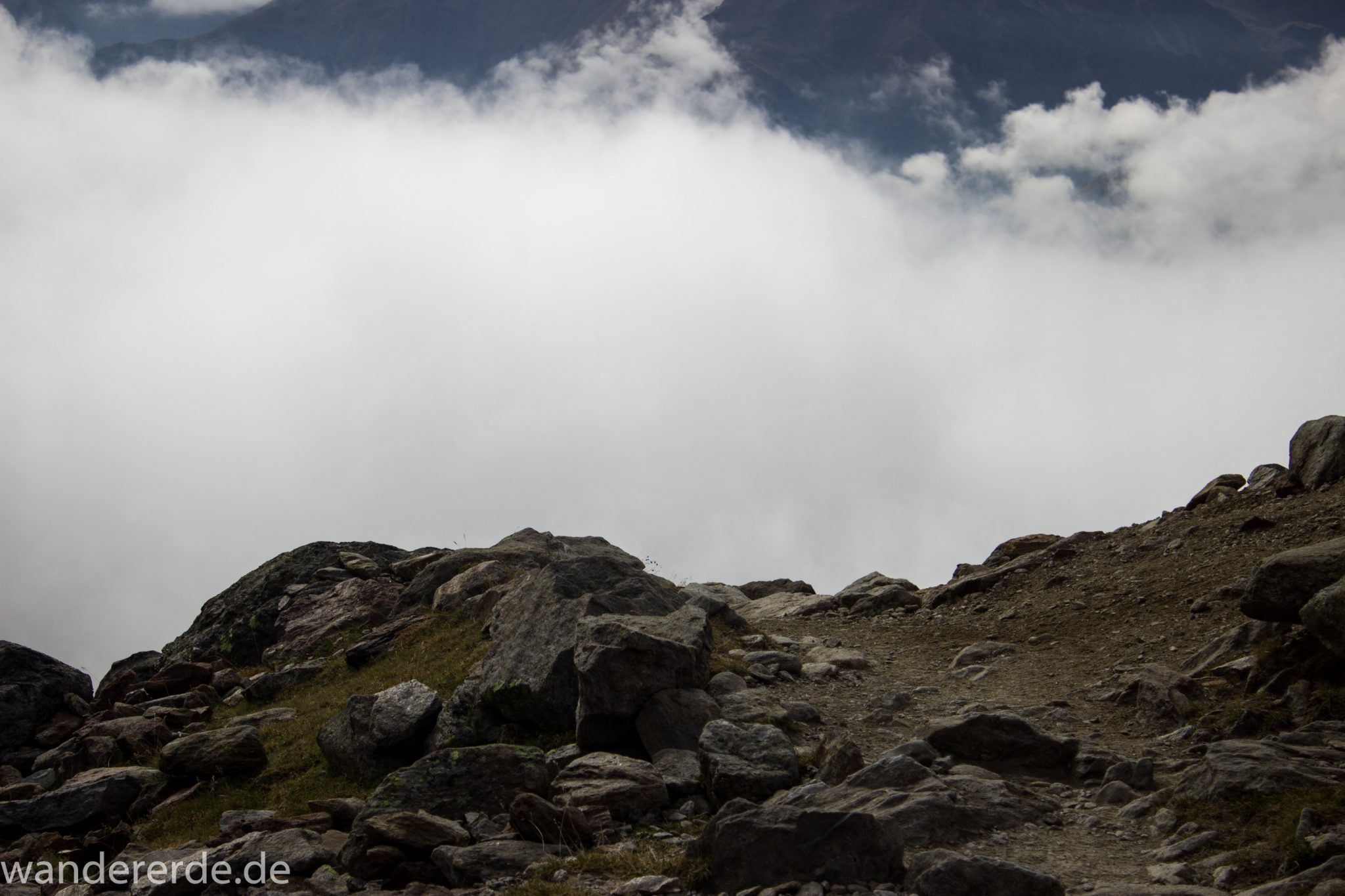 Alpenüberquerung Fernwanderweg E5 Oberstdorf Meran, 6. Etappe von Martin-Busch-Hütte nach Vernagt / Meran, nach dem Aufstieg zur Similaun-Hütte auf der letzten Etappe der Alpenüberquerung auf dem E5 verläuft der Wanderweg steil abwärts Richtung Vernagt im Schnalstal,  Bergmassiv vor einem in Wolken gehüllt, erscheint unwirklich und mystisch, Berghänge mit viel Geröll, Wanderung mit steilem Abstieg auf ausgesetztem Pfad