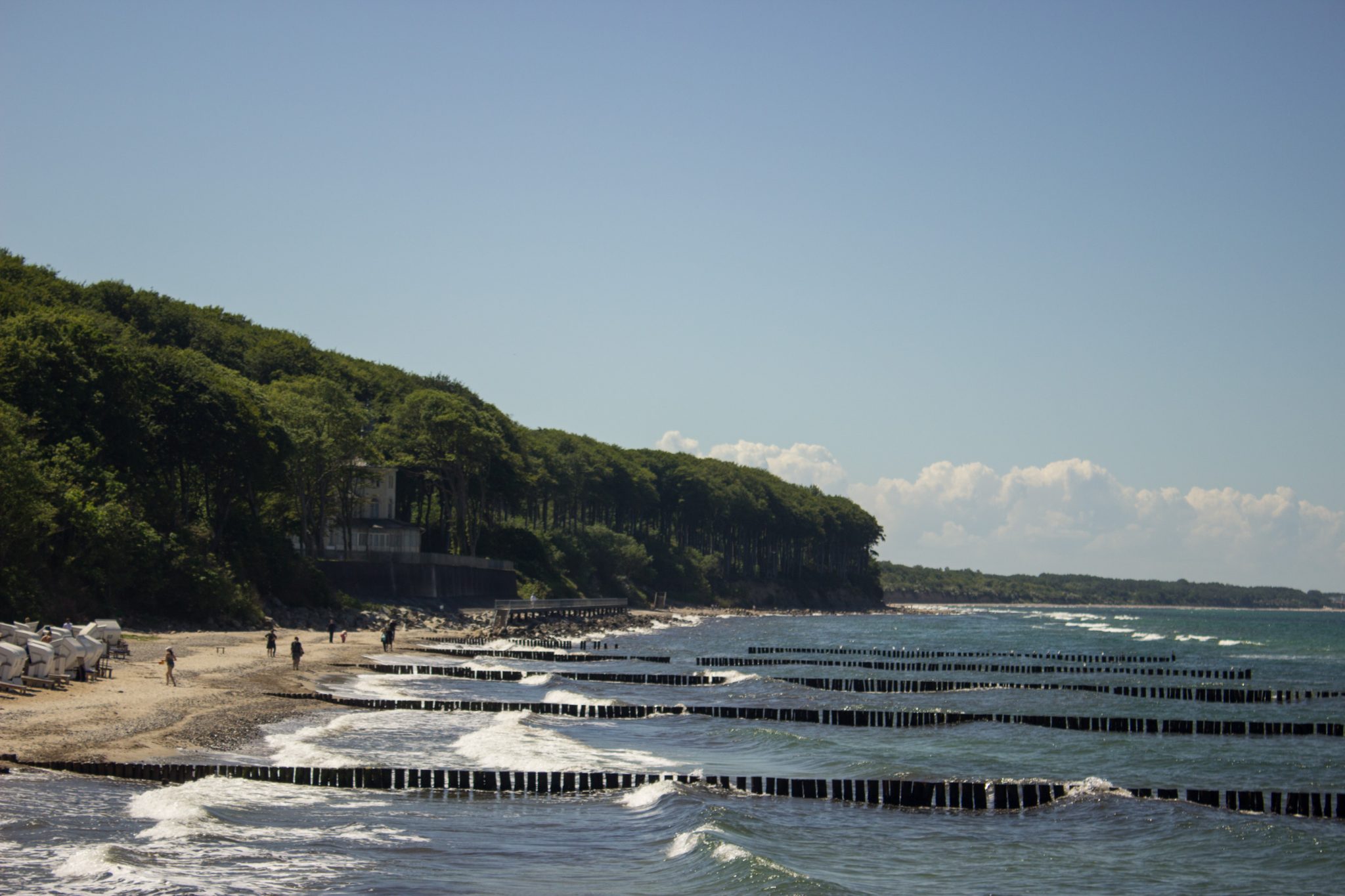 Wandern an der Ostsee von Kühlungsborn nach Börgerende auf dem E9, dritte Etappe des Wanderwegs an der Ostseeküste von Wismar nach Zingst, nach Ort Kühlungsborn führt die Wanderung weiter Richtung Heiligendamm, Blick von der Seebrücke in Heiligendamm auf die Ostsee und den dichten grünen Küstenwald, Wellen brechen am Ufer, Spaziergänger am Strand