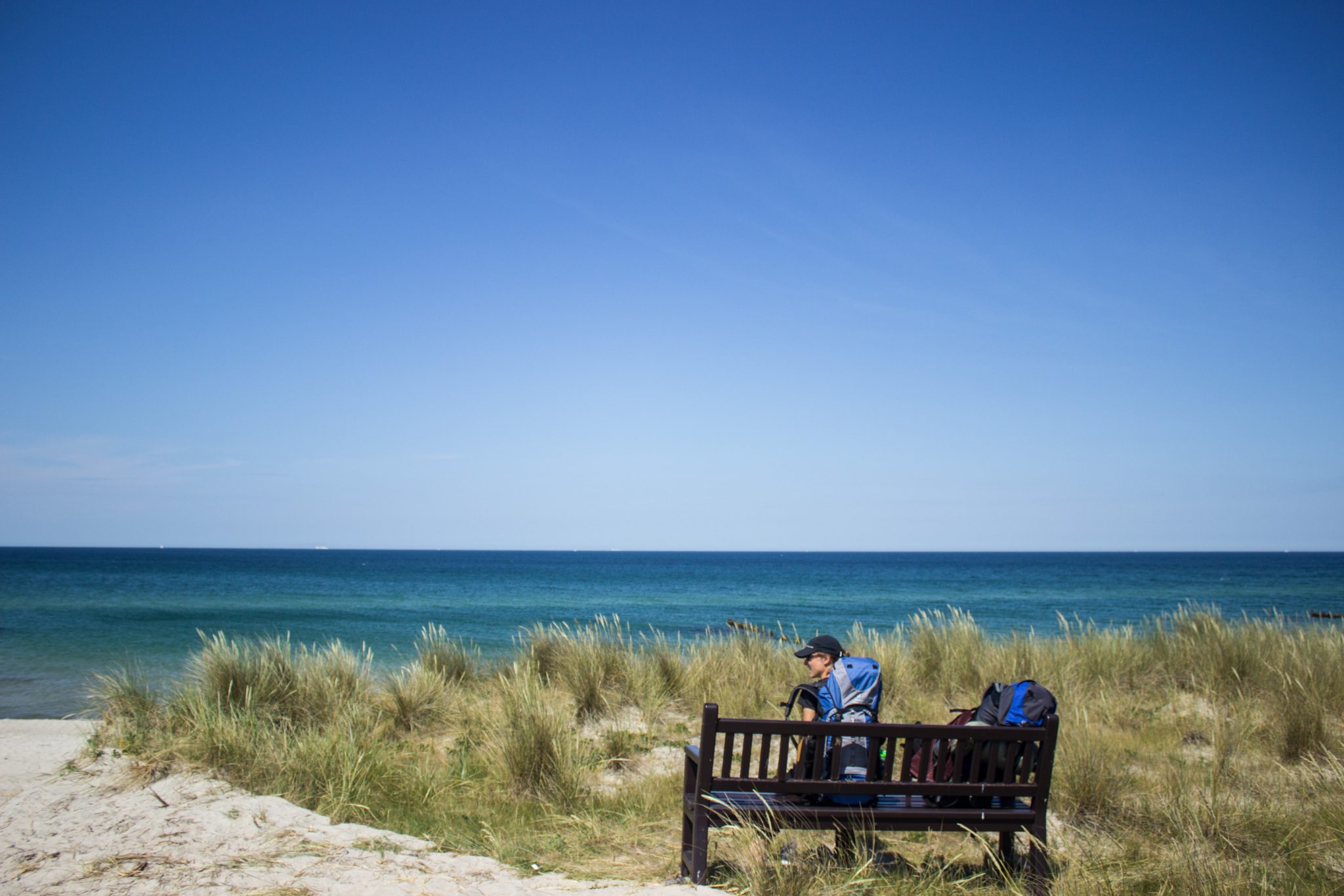 Wandern an der Ostsee von Dierhagen nach Ahrenshoop auf dem E9, sechste Etappe des Wanderwegs an der Ostseeküste von Wismar nach Zingst, da Wanderweg auf befestigter Düne verläuft entscheiden wir uns am Strand an der Ostsee weiter zu wandern, Wanderer sitzt auf Bank mit schöner Aussicht auf das Meer, wohlverdiente Pause mit Meerblick