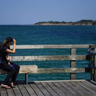 Wandern an der Ostsee von Dierhagen nach Ahrenshoop auf dem E9, sechste Etappe des Wanderwegs an der Ostseeküste von Wismar nach Zingst, am Strand an der Ostsee wandern wir bis nach Wustrow, schöne Aussicht von der Seebrücke im Ostseebad Wustrow auf die Ostsee und die Steilküste in der Ferne, Wanderer mit großem Rucksack macht Pause auf Bank, wolkenloser Himmel und Sonnenschein im Sommer