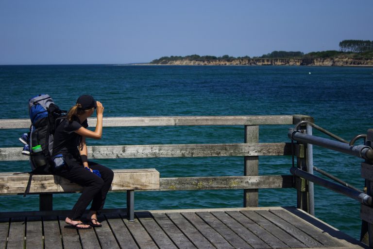 Wandern an der Ostsee von Dierhagen nach Ahrenshoop auf dem E9, sechste Etappe des Wanderwegs an der Ostseeküste von Wismar nach Zingst, am Strand an der Ostsee wandern wir bis nach Wustrow, schöne Aussicht von der Seebrücke im Ostseebad Wustrow auf die Ostsee und die Steilküste in der Ferne, Wanderer mit großem Rucksack macht Pause auf Bank, wolkenloser Himmel und Sonnenschein im Sommer