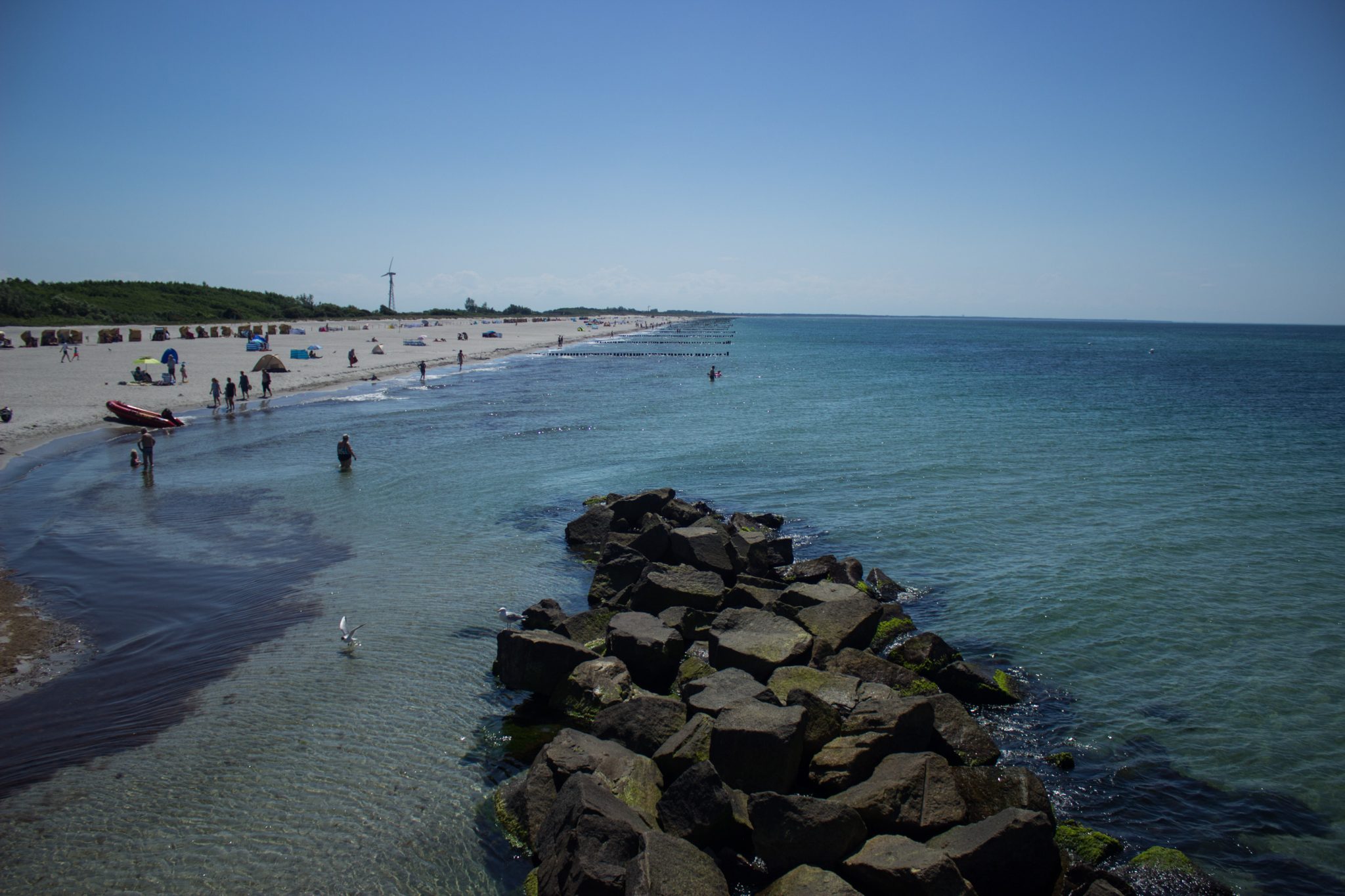 Wandern an der Ostsee von Dierhagen nach Ahrenshoop auf dem E9, sechste Etappe des Wanderwegs an der Ostseeküste von Wismar nach Zingst, am Strand an der Ostsee wandern wir bis zum Ostseebad Wustrow, traumhaft schöne Aussicht auf das Meer mit Mole und den Strand, Ostsee mit sauberem klarem Wasser, kleine Wellen brechen am Ufer, Spaziergänger und Urlauber am Strand, wolkenloser Himmel und Sonnenschein im Sommer