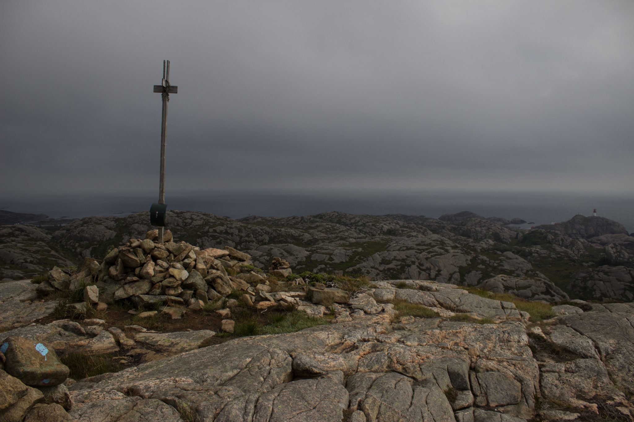 Wanderungen beim Kap Lindesnes, südlichster Punkt Norwegens, am Südkap in Norwegen, Wanderer unterwegs auf schmalem Pfad entlang großer Felsen, kleiner Berggipfel mit Gipfelkreuz, grüne Vegetation an der Küste, Aussicht auf das weite Meer und den Leuchtturm Lindesnes Fyr