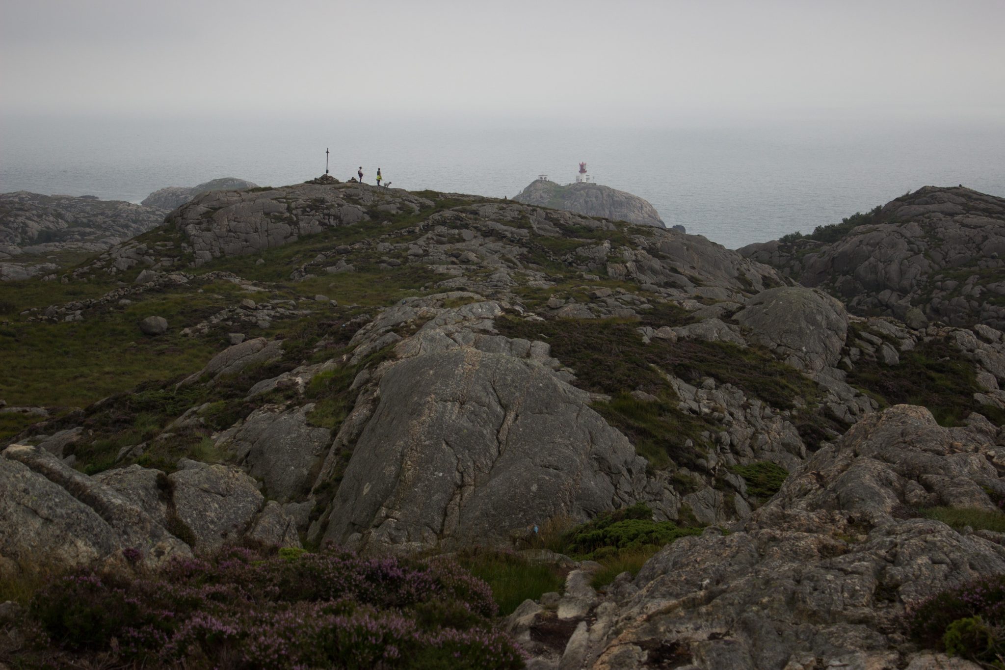 Wanderungen beim Kap Lindesnes, südlichster Punkt Norwegens, am Südkap in Norwegen, Wanderer unterwegs auf schmalem Pfad entlang großer Felsen, kleiner Berggipfel mit Gipfelkreuz, grüne Vegetation an der Küste, Aussicht auf das weite Meer und den Leuchtturm Lindesnes Fyr