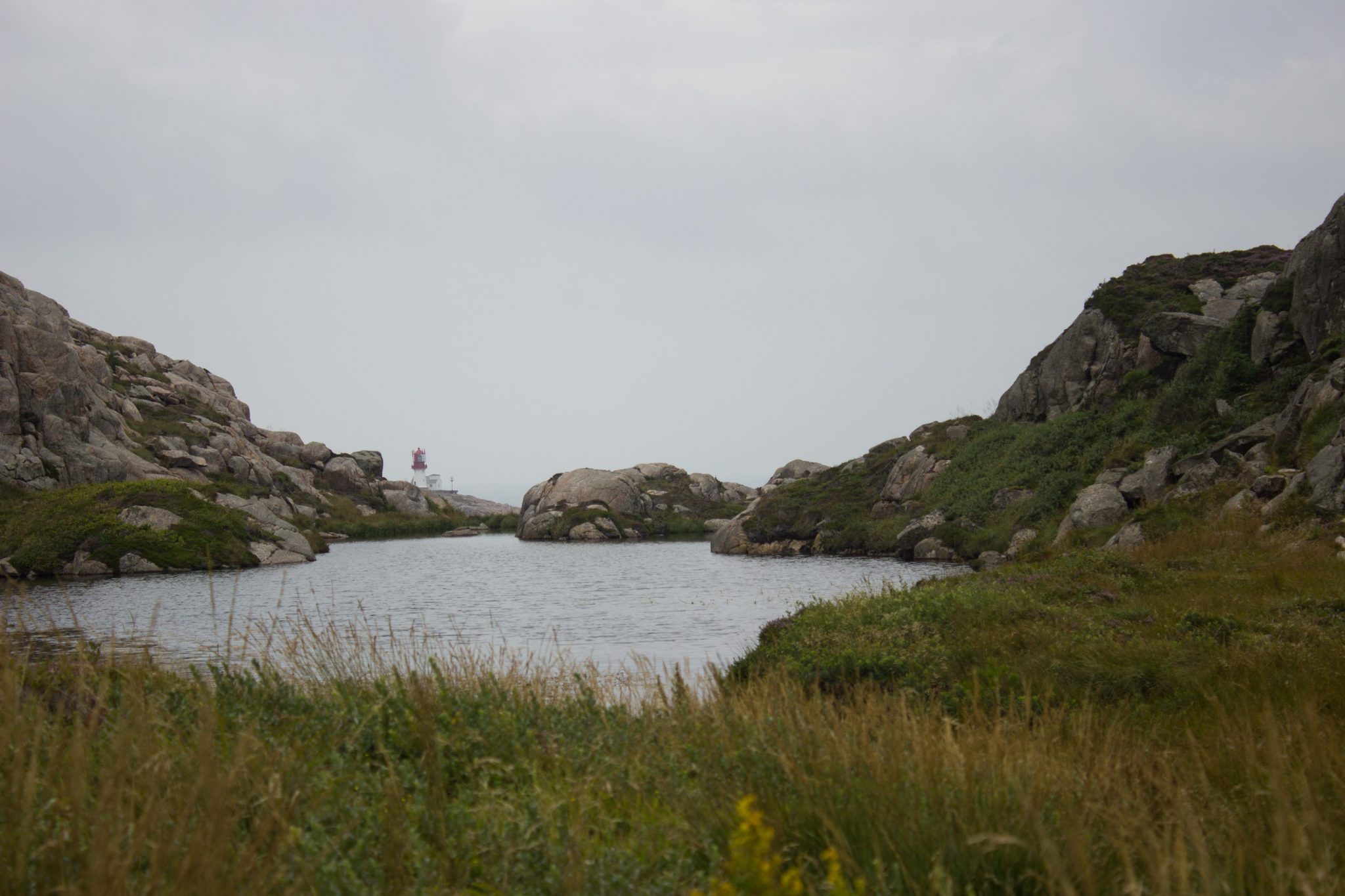 Wanderungen beim Kap Lindesnes, südlichster Punkt Norwegens, am Südkap in Norwegen, Wanderer unterwegs auf schmalem Pfad entlang großer Felsen, grüne Vegetation an der Küste, Heidelandschaft, Aussicht auf das weite Meer und einem kleinen See, im Hintergrund der Leuchtturm Lindesnes Fyr