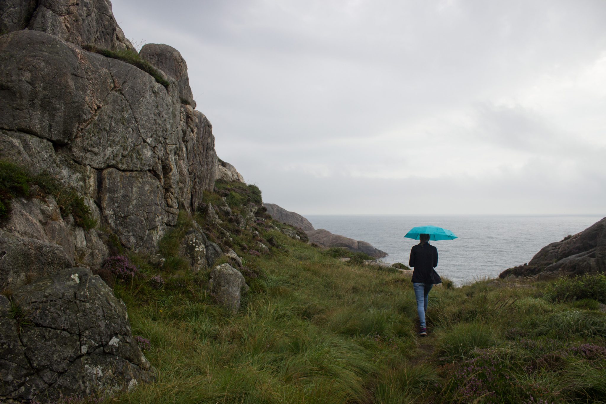 Wanderungen beim Kap Lindesnes, südlichster Punkt Norwegens, am Südkap in Norwegen, Wanderer im Regen unterwegs auf schmalem Pfad entlang großer Felsen, teils sehr sumpfiger Weg, grüne Vegetation an der Küste, Heidelandschaft, Aussicht auf das weite Meer