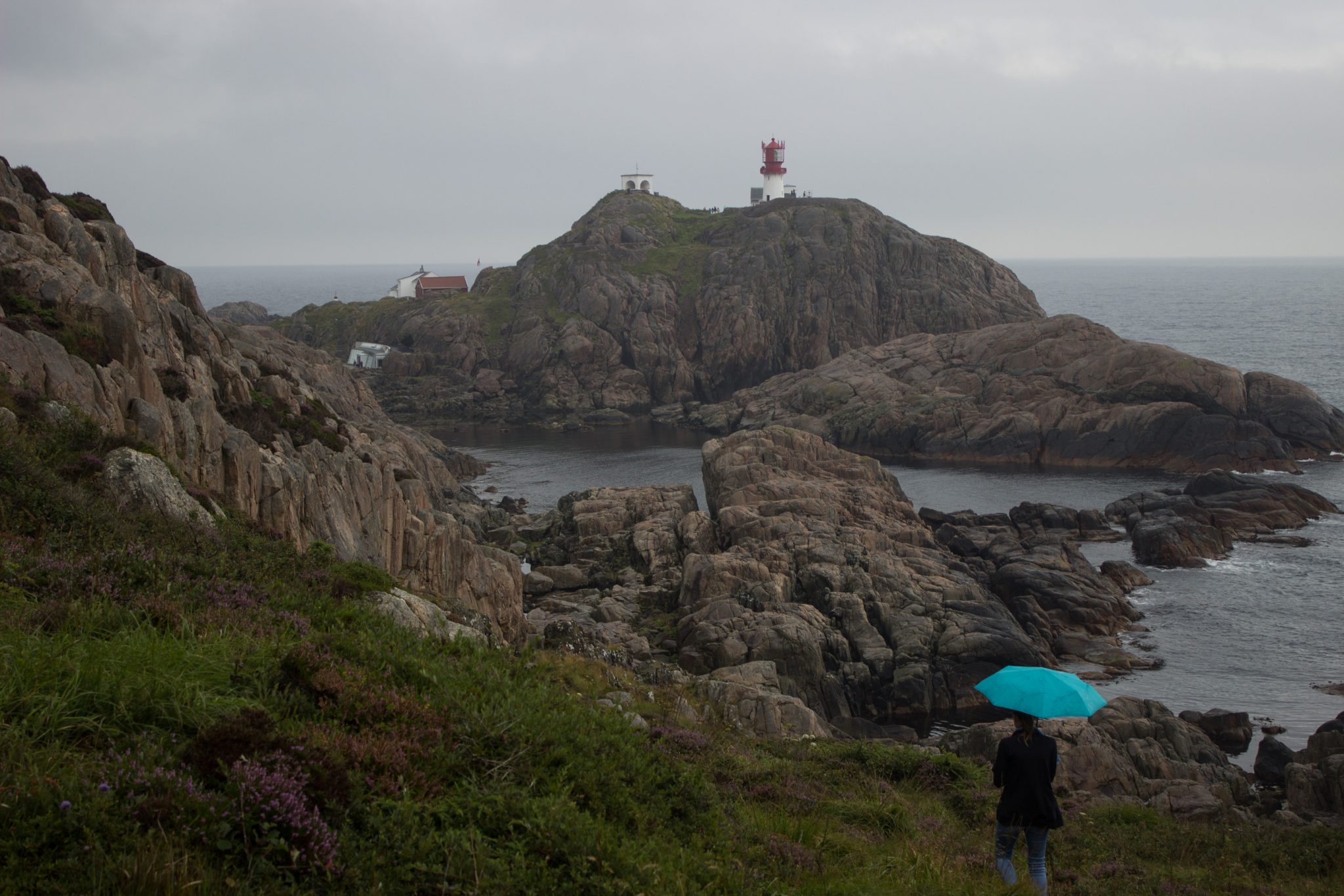 Wanderungen beim Kap Lindesnes, südlichster Punkt Norwegens, am Südkap in Norwegen, Wanderer im Regen unterwegs auf schmalem Pfad entlang großer Felsen, teils sehr sumpfiger Weg, grüne Vegetation an der Küste, Heidelandschaft, Aussicht auf das weite Meer und den Leuchtturm Lindesnes Fyr