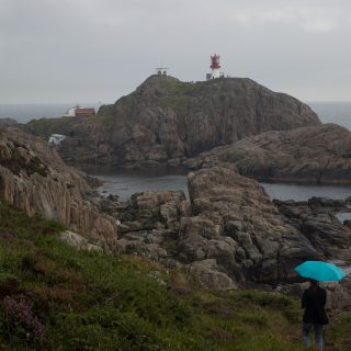 Wanderungen beim Kap Lindesnes, südlichster Punkt Norwegens, am Südkap in Norwegen, Wanderer im Regen unterwegs auf schmalem Pfad entlang großer Felsen, teils sehr sumpfiger Weg, grüne Vegetation an der Küste, Heidelandschaft, Aussicht auf das weite Meer und den Leuchtturm Lindesnes Fyr