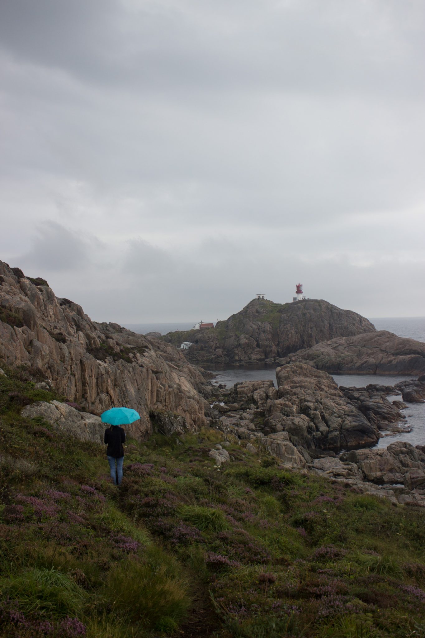 Wanderungen beim Kap Lindesnes, südlichster Punkt Norwegens, am Südkap in Norwegen, Wanderer im Regen unterwegs auf schmalem Pfad entlang großer Felsen, teils sehr sumpfiger Weg, grüne Vegetation an der Küste, Heidelandschaft, Aussicht auf das weite Meer und den Leuchtturm Lindesnes Fyr