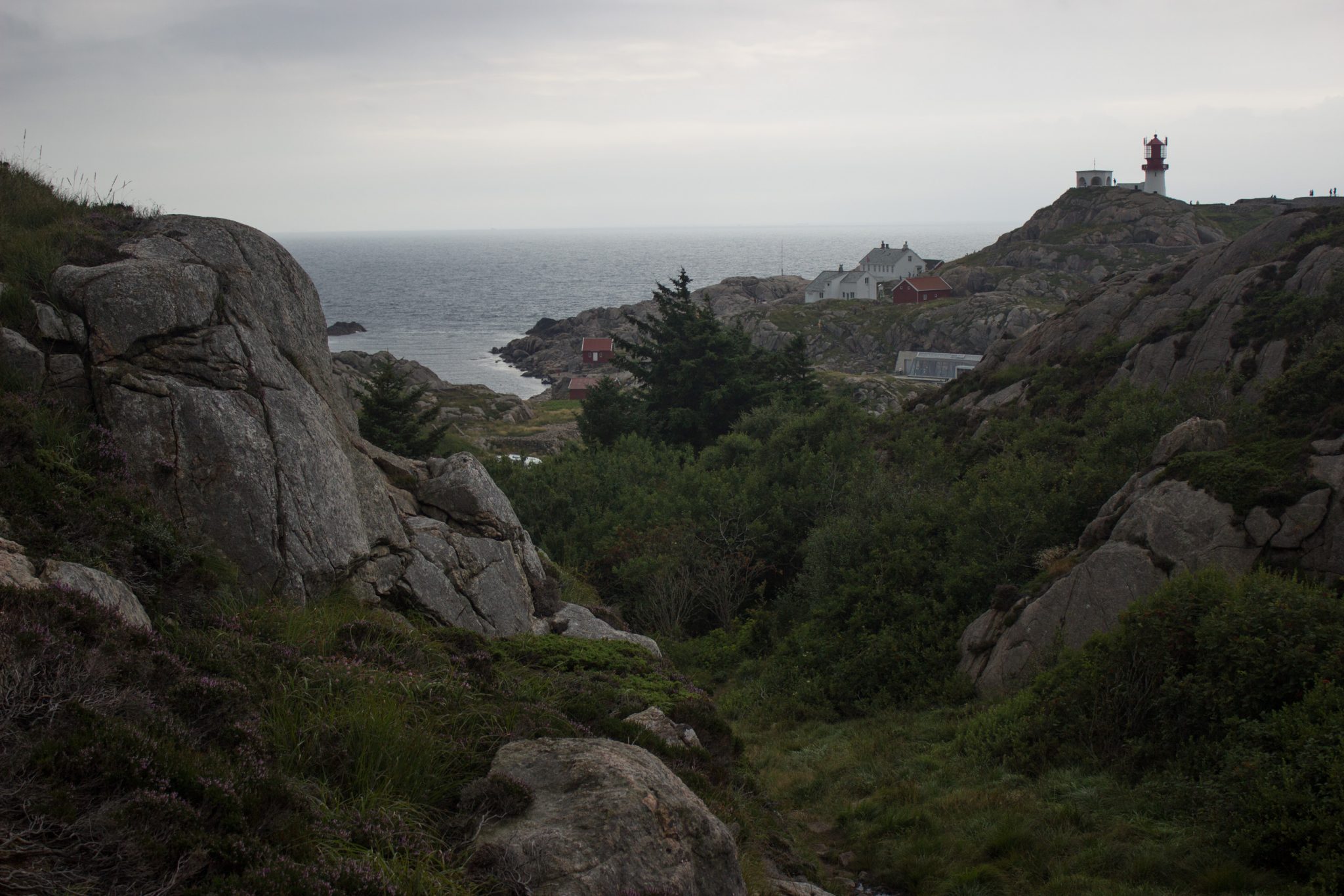 Wanderungen beim Kap Lindesnes, südlichster Punkt Norwegens, am Südkap in Norwegen, während der Wanderung Aussicht auf den Leuchtturm und ein paar Häuser, dahinter das weite Meer, Wanderweg führt über große Felsen, grüne Vegetation an der Küste