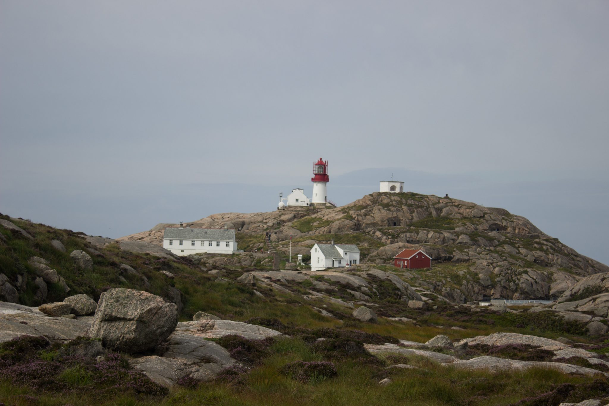 Wanderungen beim Kap Lindesnes, südlichster Punkt Norwegens, Leuchtturm Lindesnes Fyr am Südkap in Norwegen, Wanderer unterwegs auf schmalem Pfad entlang und über große Felsen, grüne Vegetation an der Küste, Heidelandschaft, Aussicht auf den Leuchtturm Lindesnes Fyr und das dahinterliegende weite Meer